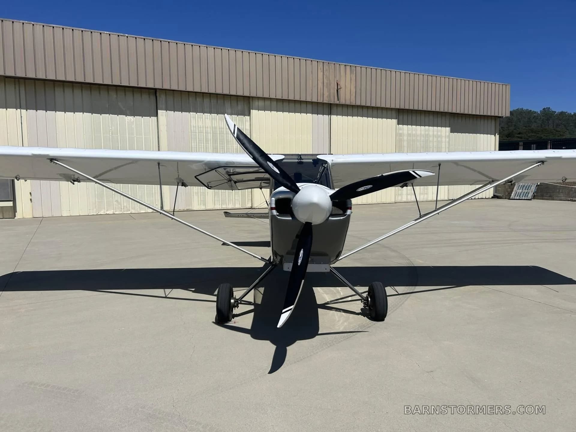 A head-on view of a white and grey Rans S-21 Outbound aircraft with a large propeller, parked in front of a hangar.