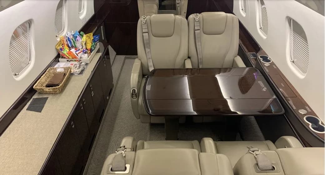 A section of the aircraft cabin interior with a large table, beige leather seats, and a galley counter with snacks.