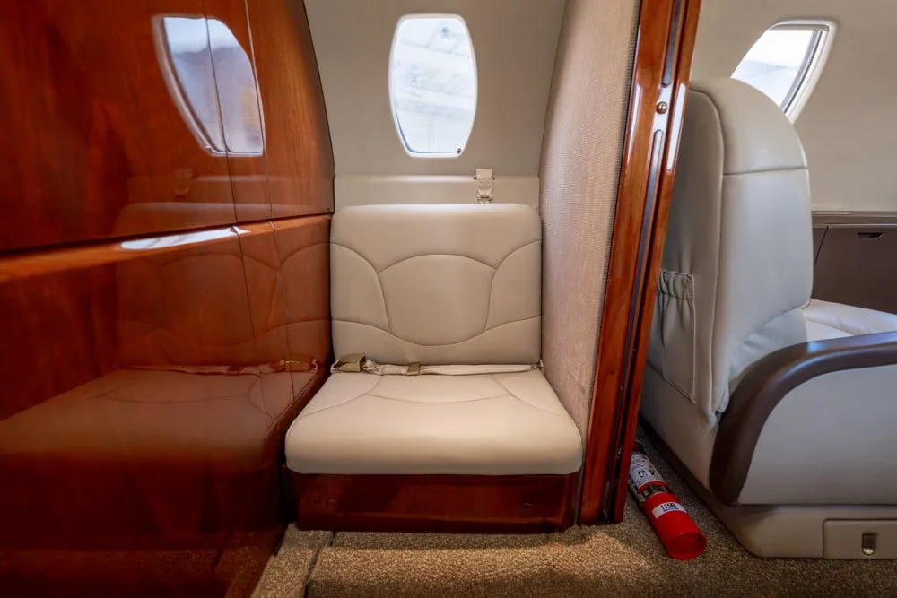 A single beige leather seat in the private jet cabin, positioned next to a wooden partition and a red fire extinguisher.