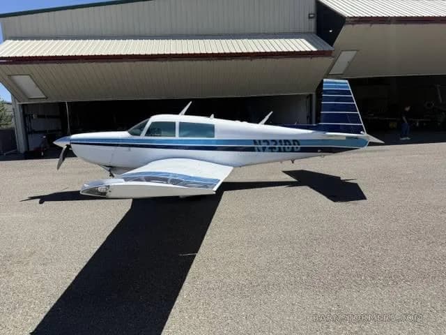 A white and blue Mooney aircraft, registration N231DD, parked on an asphalt ramp in front of an open hangar.