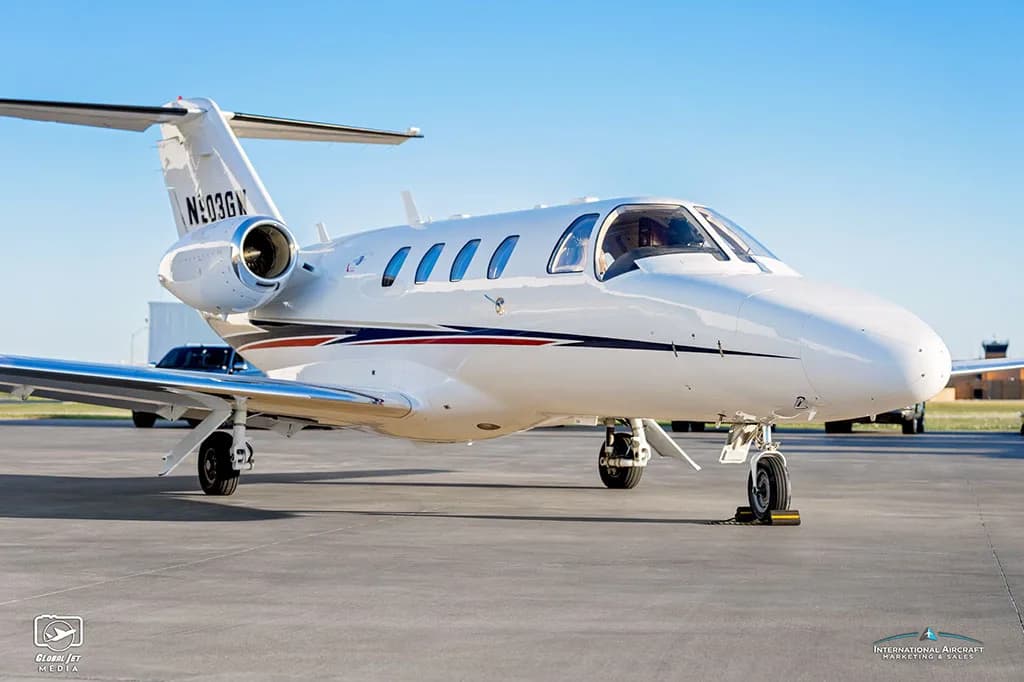 A front-side view of a white Cessna Citation CJ3 (N903GW) with blue and red stripes parked on an airfield.