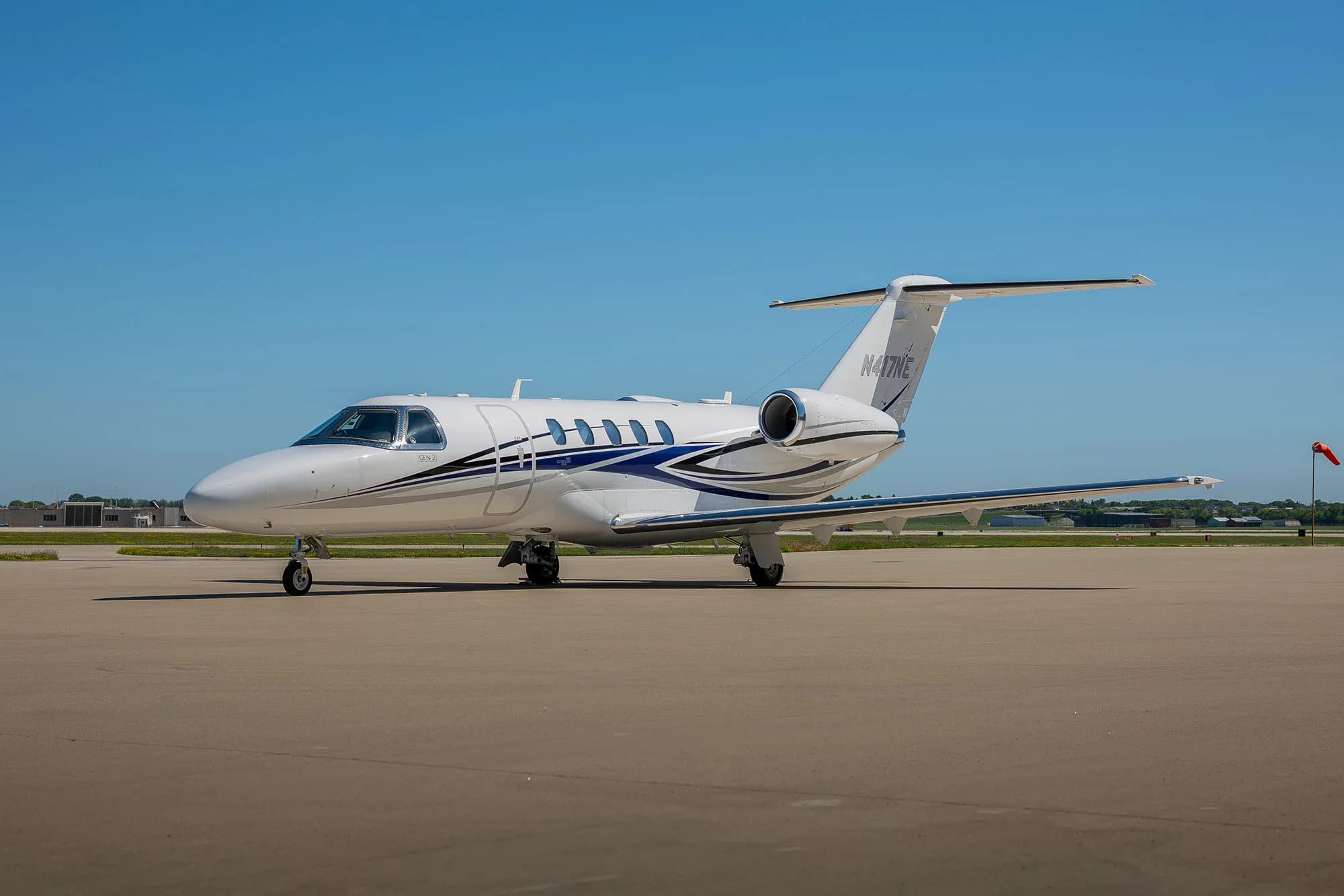 A white business jet with blue and grey stripes parked on an airfield with a red windsock in the background.