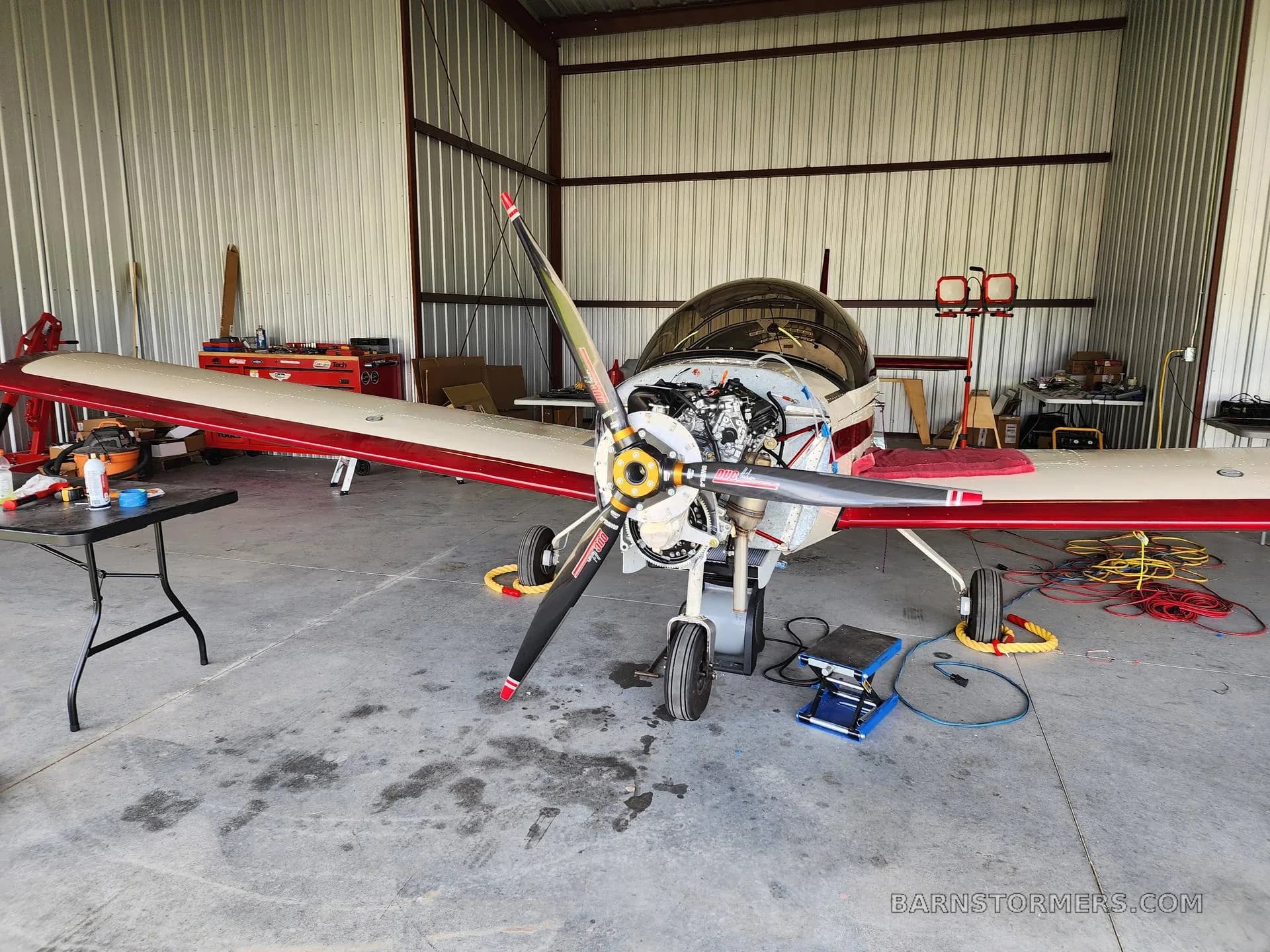 A white and red single-engine aircraft is in a hangar with its engine cowling removed for maintenance.