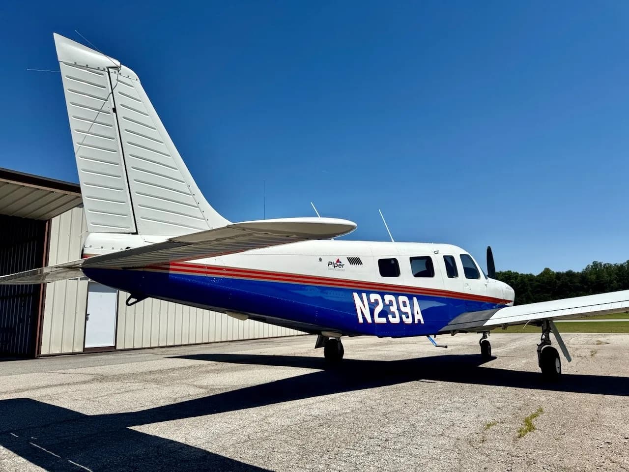 A side view of a white and blue Piper Saratoga II HP aircraft with red stripes, registration N239A, parked on an airfield.