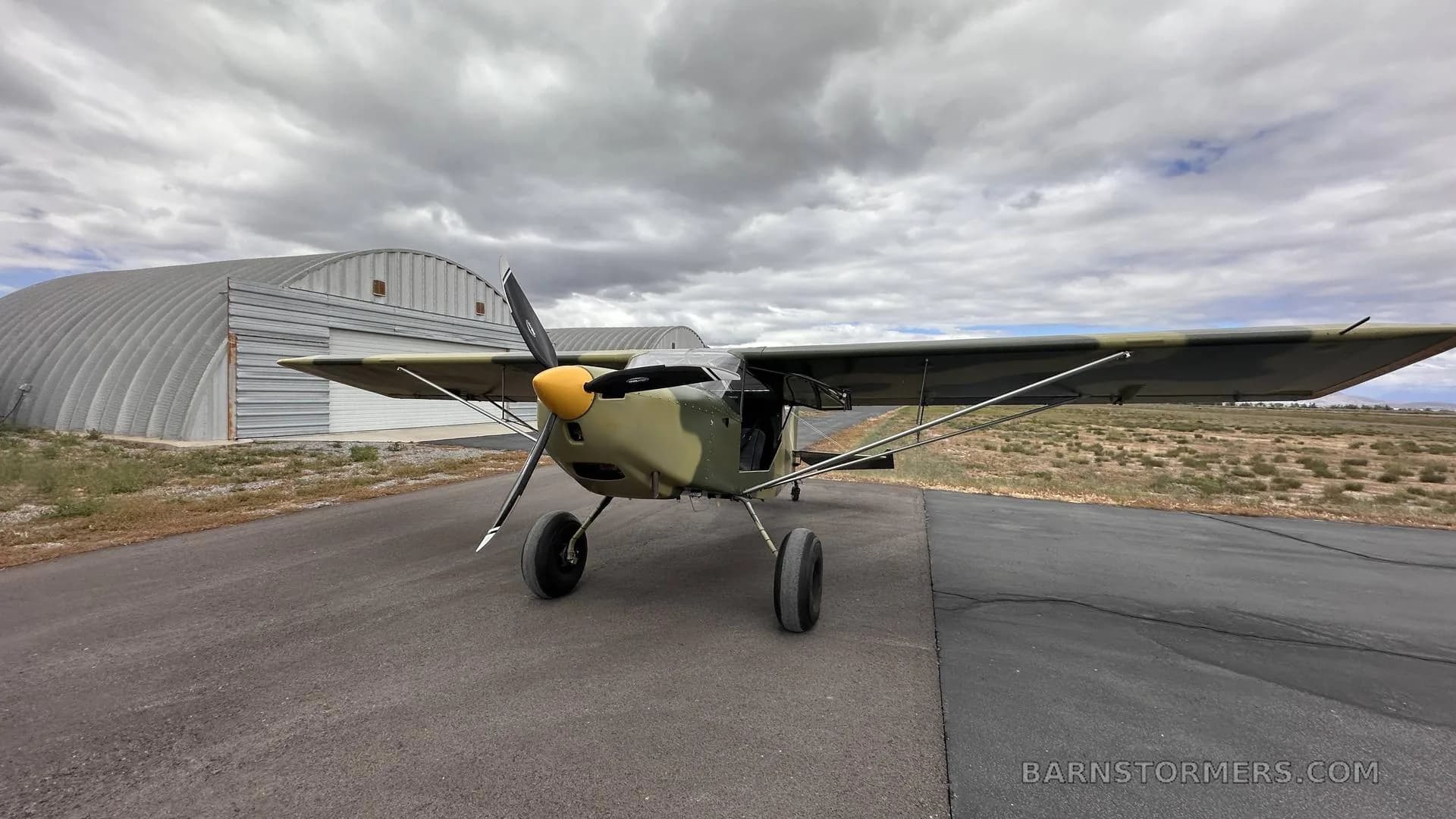 A light aircraft with camouflage paint and a yellow propeller spinner is parked on an asphalt runway in front of hangars under a cloudy sky.