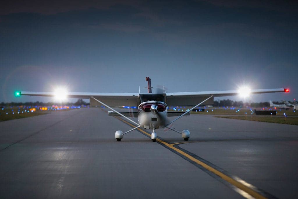 A white and maroon Cessna 182 aircraft with its landing lights on, positioned on a runway at dusk.