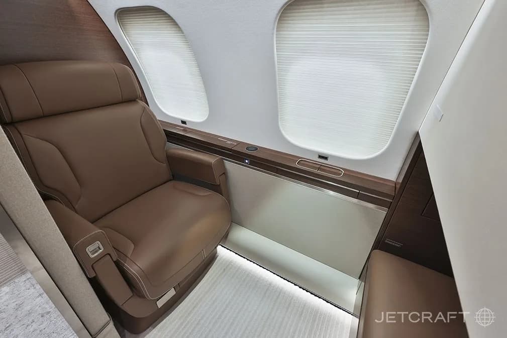 A luxurious brown leather single seat in the cabin of a business jet, next to two windows with shades.