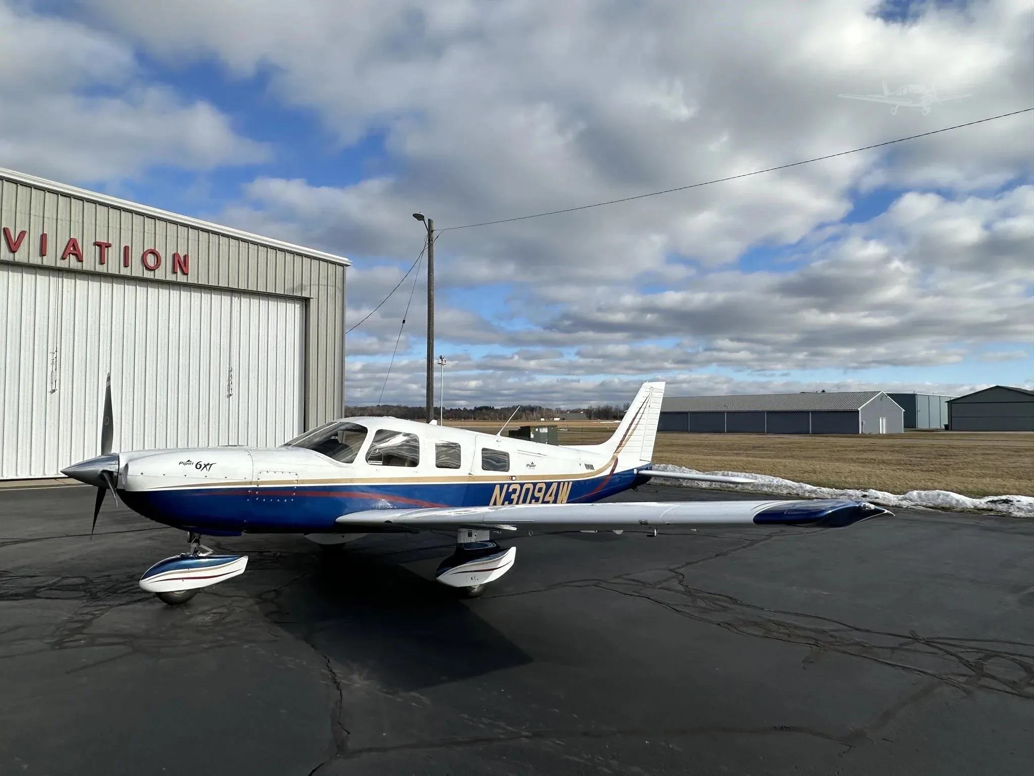 A side view of a blue and white Piper Malibu Mirage N3094W parked on an asphalt ramp in front of a Bartelt Aviation hangar.