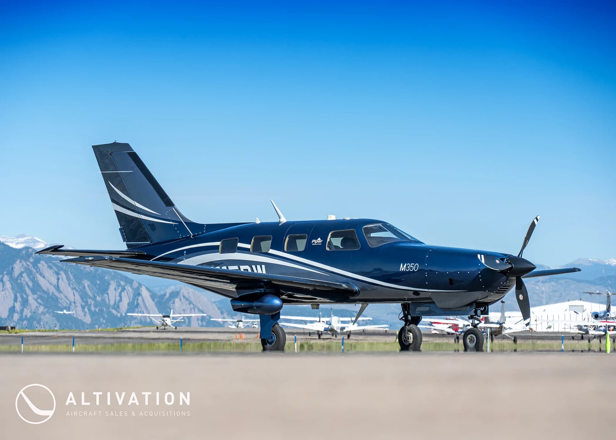 A dark blue Piper M350 aircraft with white and grey accents parked on an airfield with mountains in the background.