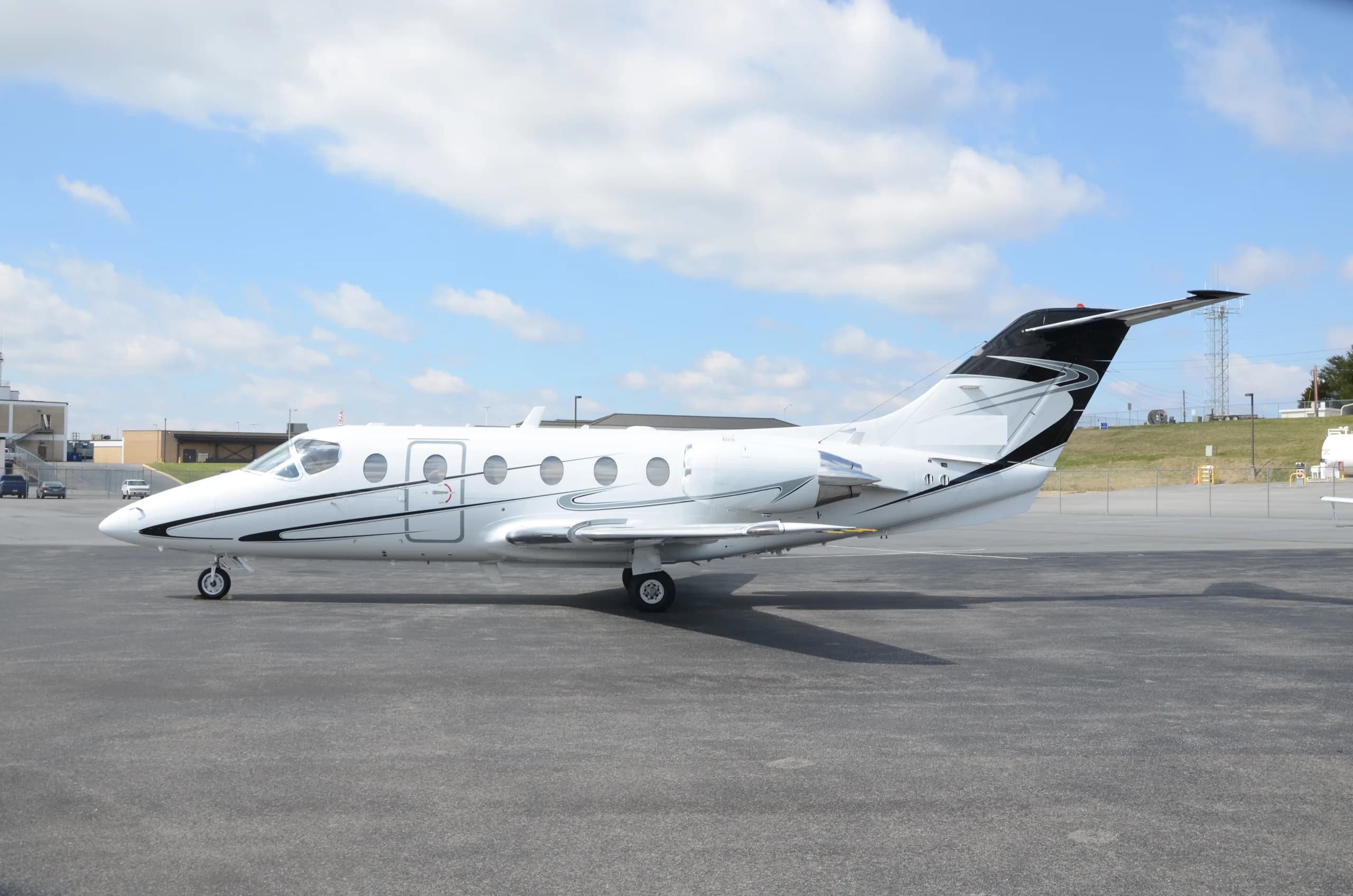 A white business jet with black and silver accent stripes parked on an asphalt ramp under a partly cloudy sky.