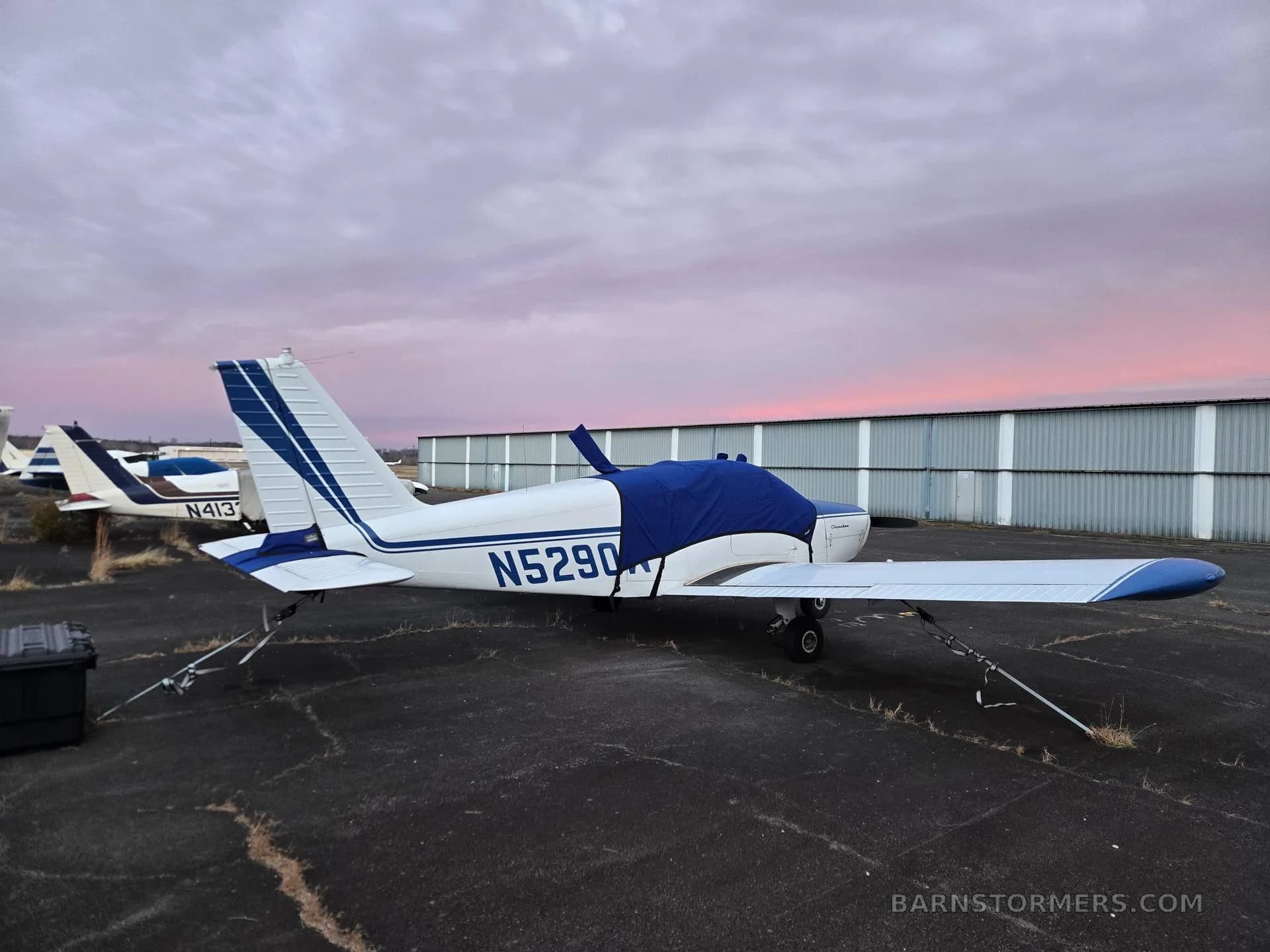 A white and blue single-engine aircraft (N5290W) with a blue cover, parked on an airfield at sunset.