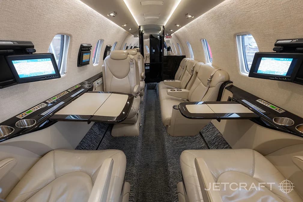 The interior cabin of a business jet with light beige leather seats, fold-out tables, and individual entertainment screens.
