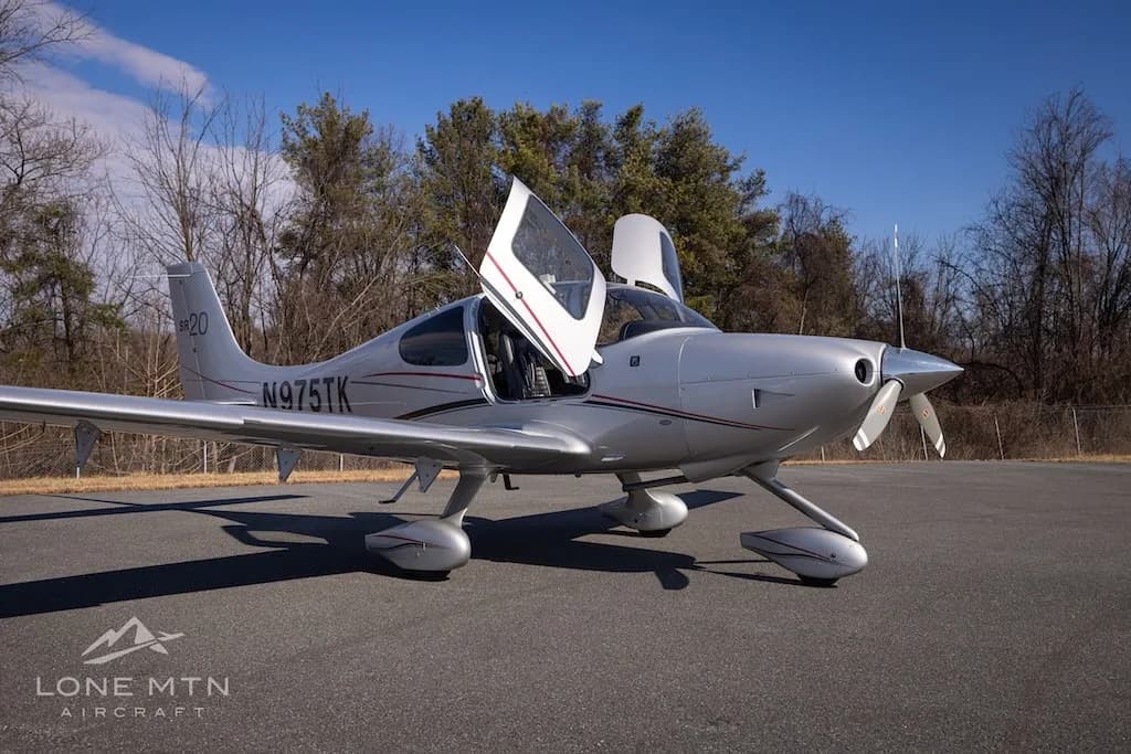A silver Cirrus SR20 aircraft with registration N975TK parked on an asphalt runway, viewed from the front-right with doors open.