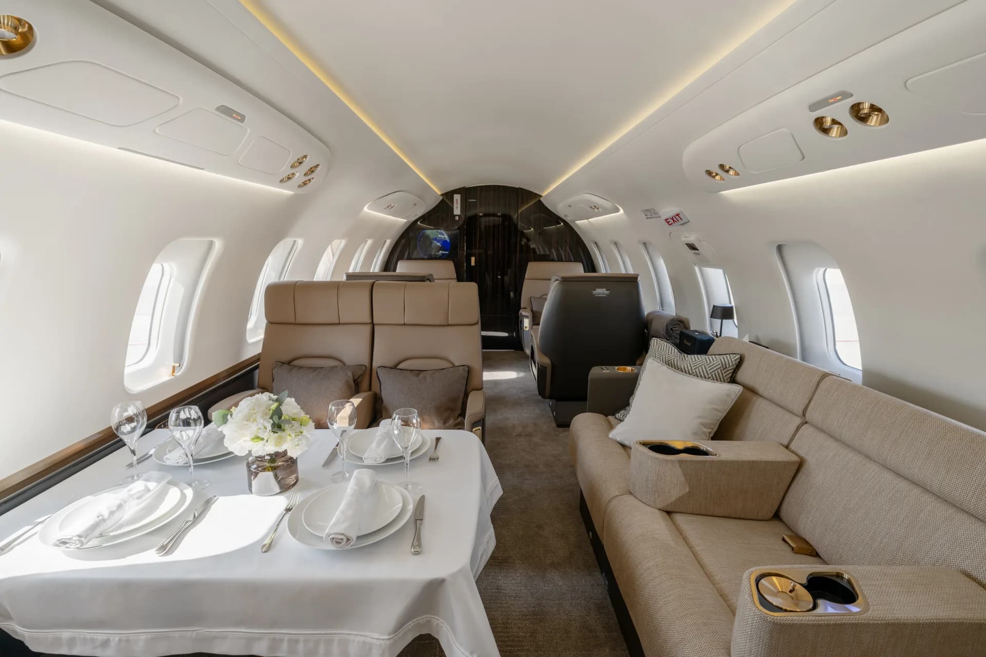 A spacious aircraft cabin interior with a dining table on the left and a long beige sofa on the right, looking towards the rear.