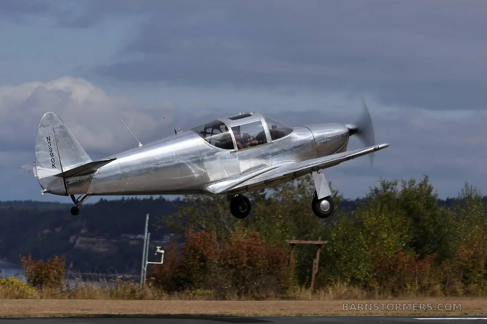 A shiny silver single-engine aircraft with tail number N3390K is captured in flight, just above a runway with trees and water in the background.