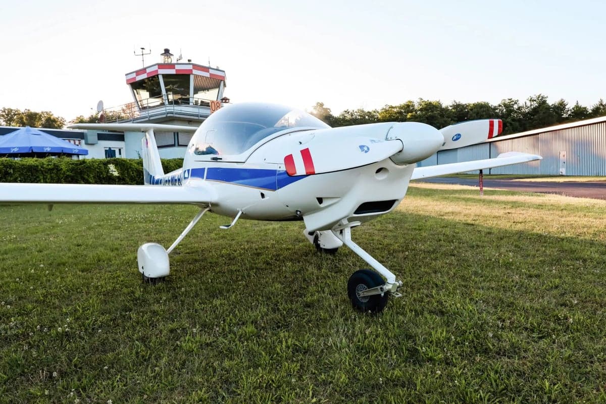 Diamond DA20 Katana D-EMFH parked on grass with an airport control tower in the background.