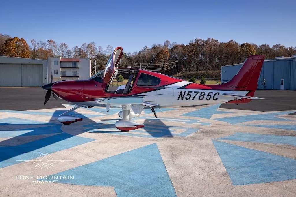 A red and white Cirrus SR22 aircraft with registration N5785C parked on an airfield with its doors open, buildings in the background.