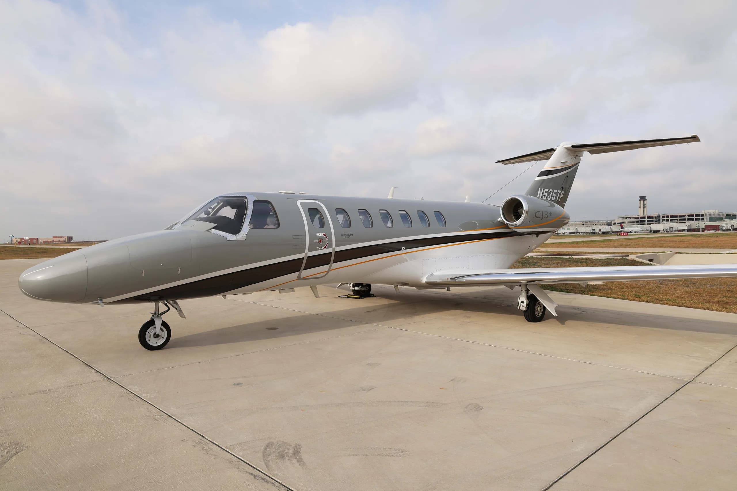 A Cessna Citation CJ3+ business jet, registration N535TP, with a grey and white paint scheme and dark brown and gold accent stripes, parked on an airfield under a cloudy sky.