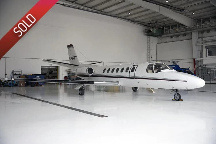 A white Cessna Citation business jet with a dark stripe is shown from the side in a brightly lit hangar.