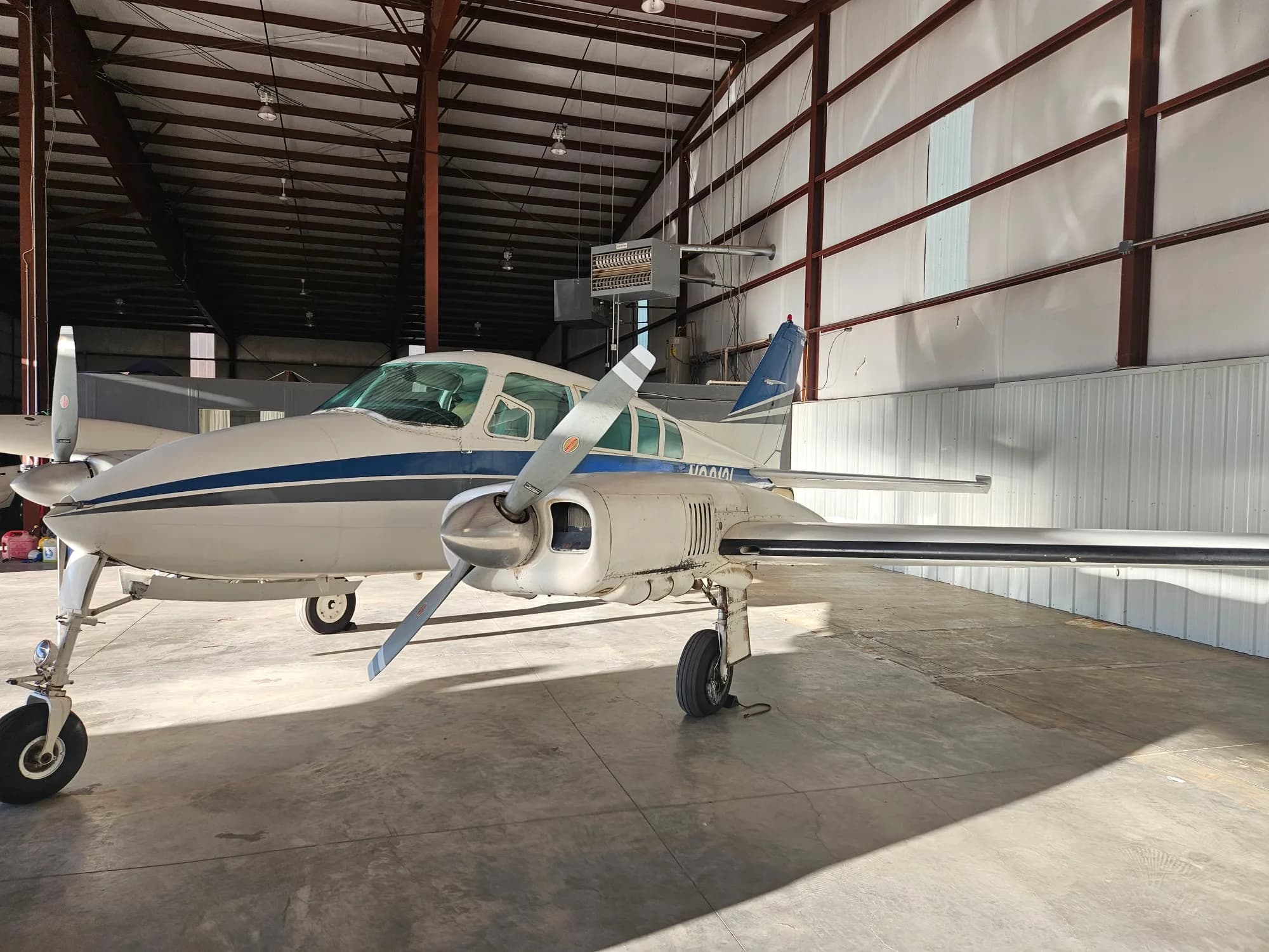 A white twin-engine propeller aircraft with a blue stripe is parked inside a hangar, viewed from the front-left.