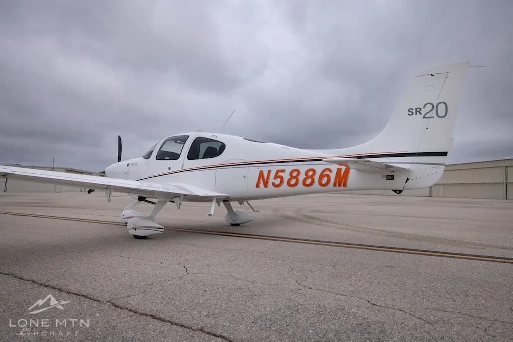 A side view of a white Cirrus SR20 aircraft with orange and black stripes, registration N5886M, on an airfield.
