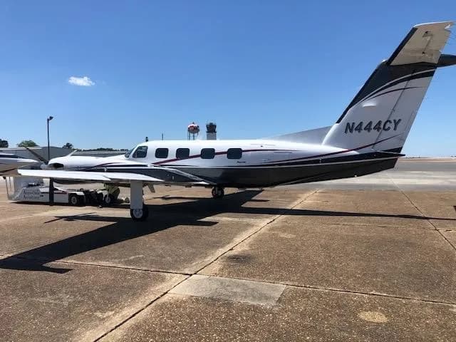 A white and dark grey twin-engine turboprop aircraft with red pinstripes, tail number N444CY, is parked on an airport tarmac with an air traffic control tower in the background.