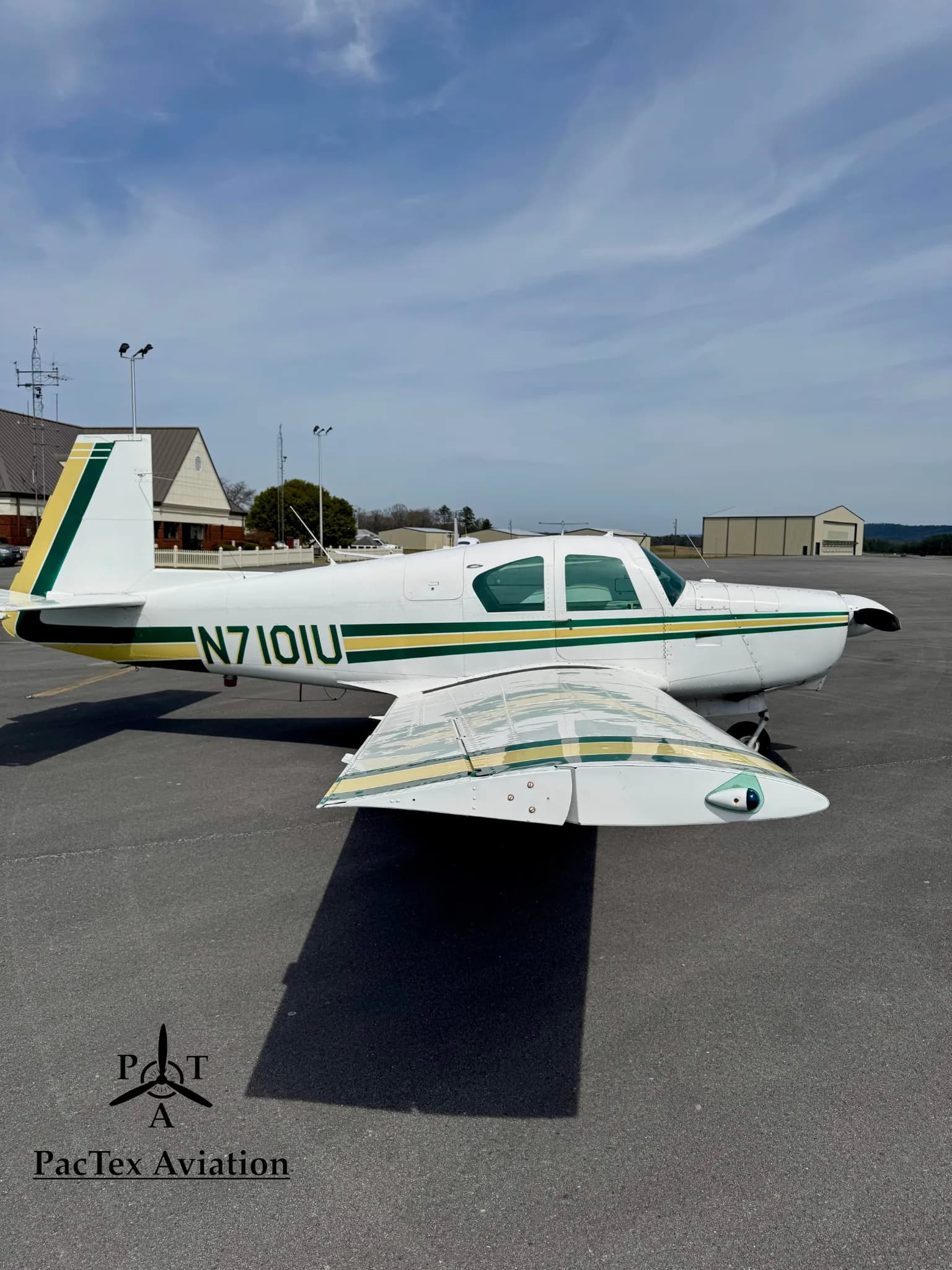 A white Mooney M20C aircraft with green and yellow stripes, parked on an airport tarmac, viewed from the side.