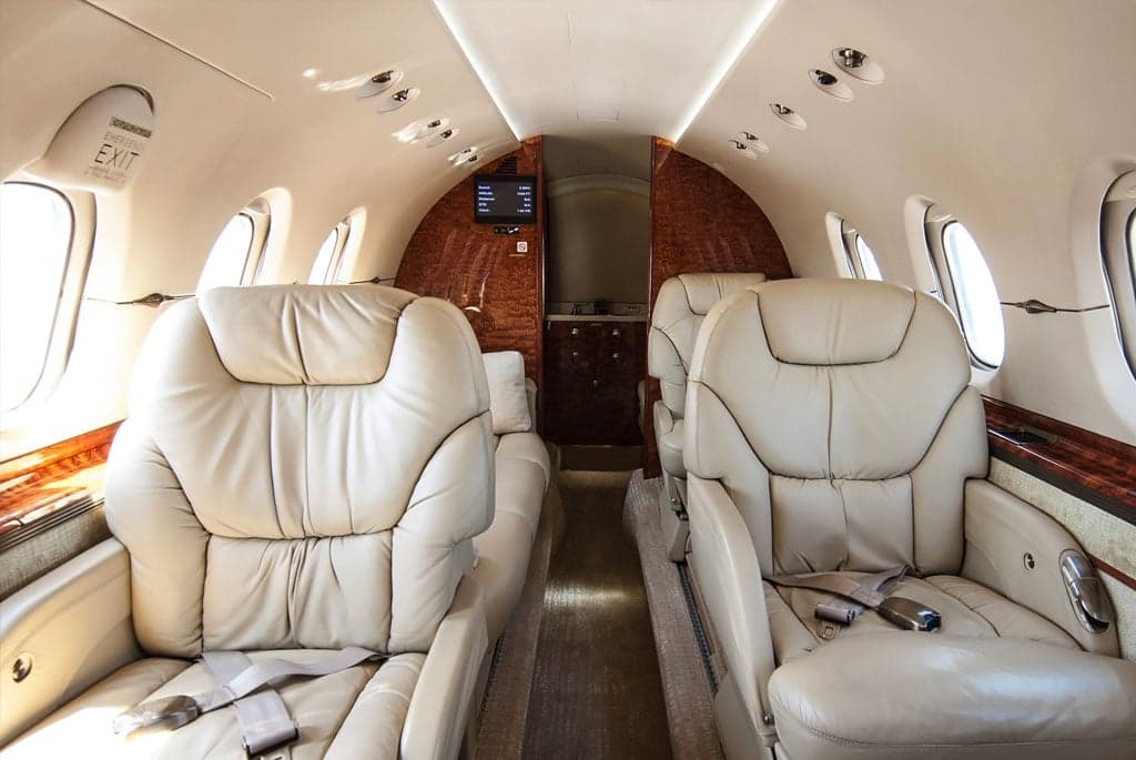 A perspective view down the aisle of the business jet's cabin, showing facing cream leather seats and wood paneling.