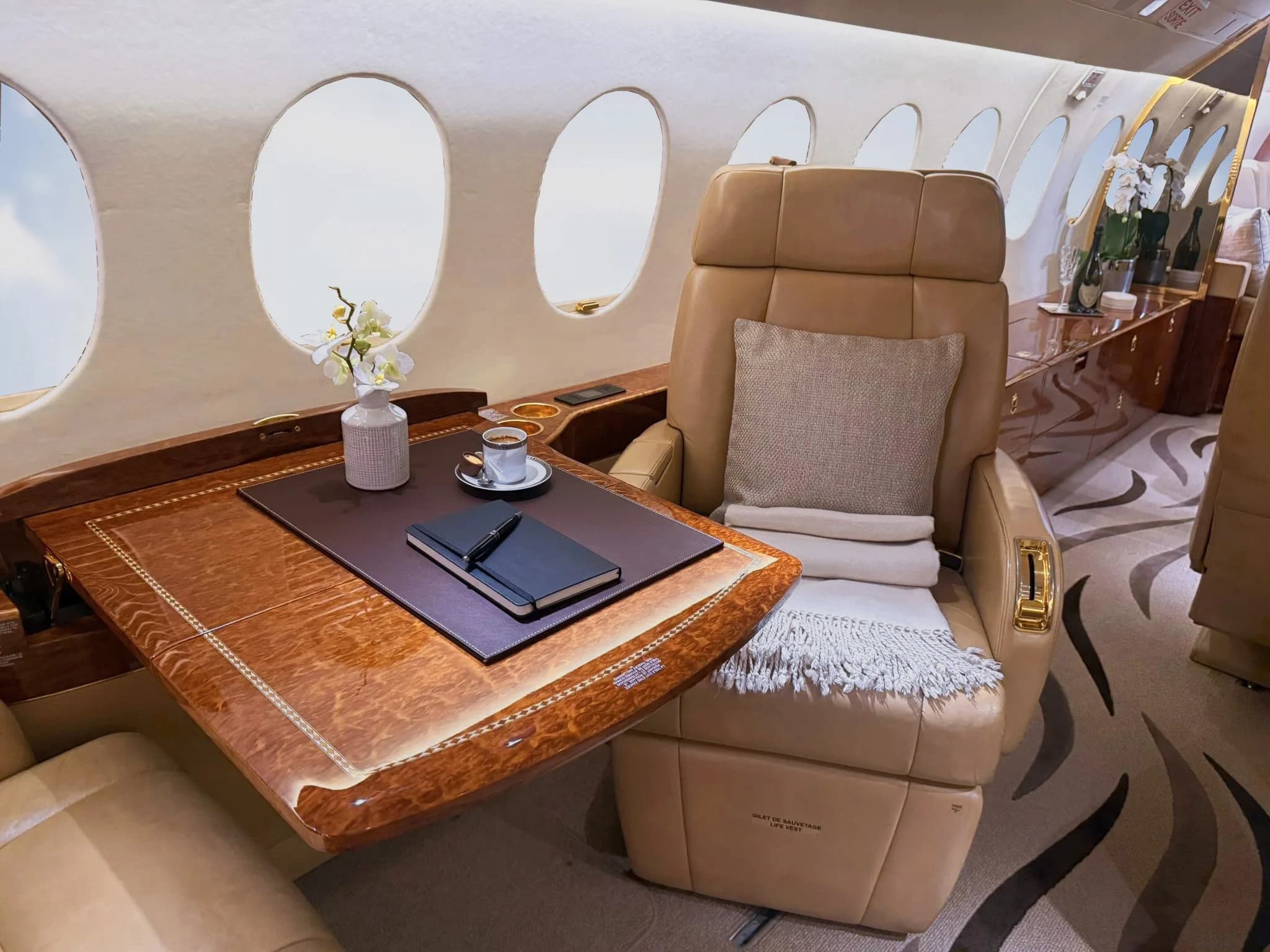 A close-up view of a private jet's interior shows a tan leather seat next to a polished wooden table with a notebook and coffee cup.