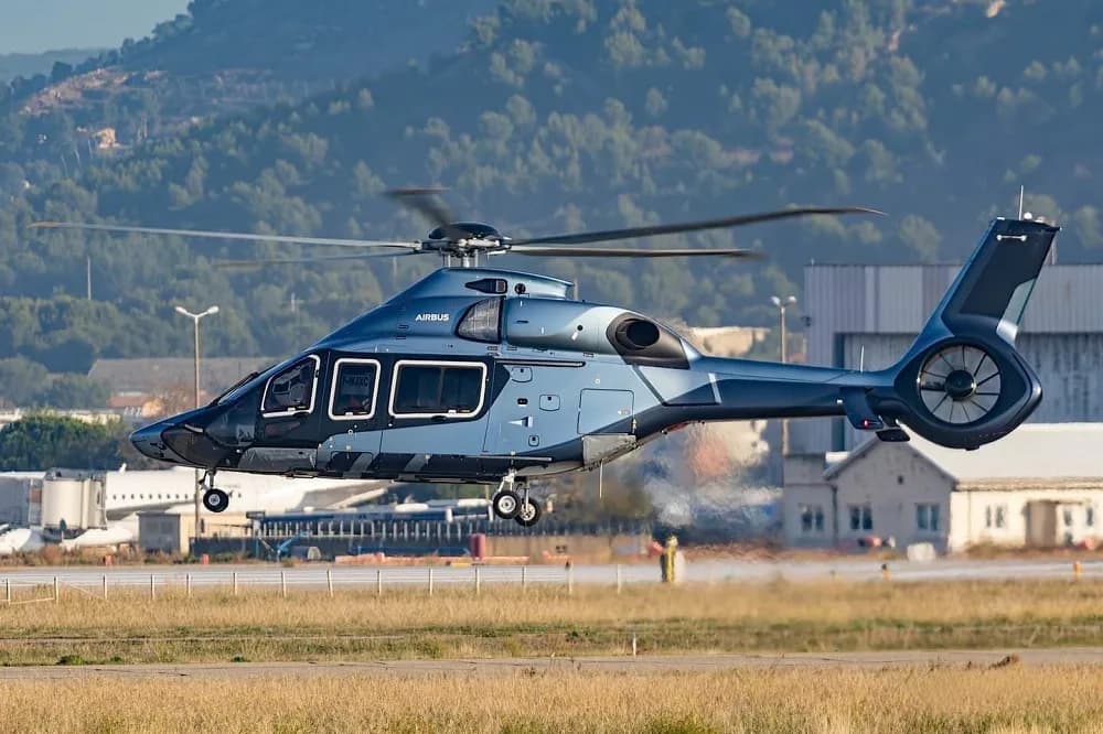 A blue and grey Airbus H175 helicopter hovering above an airport runway with mountains in the background.