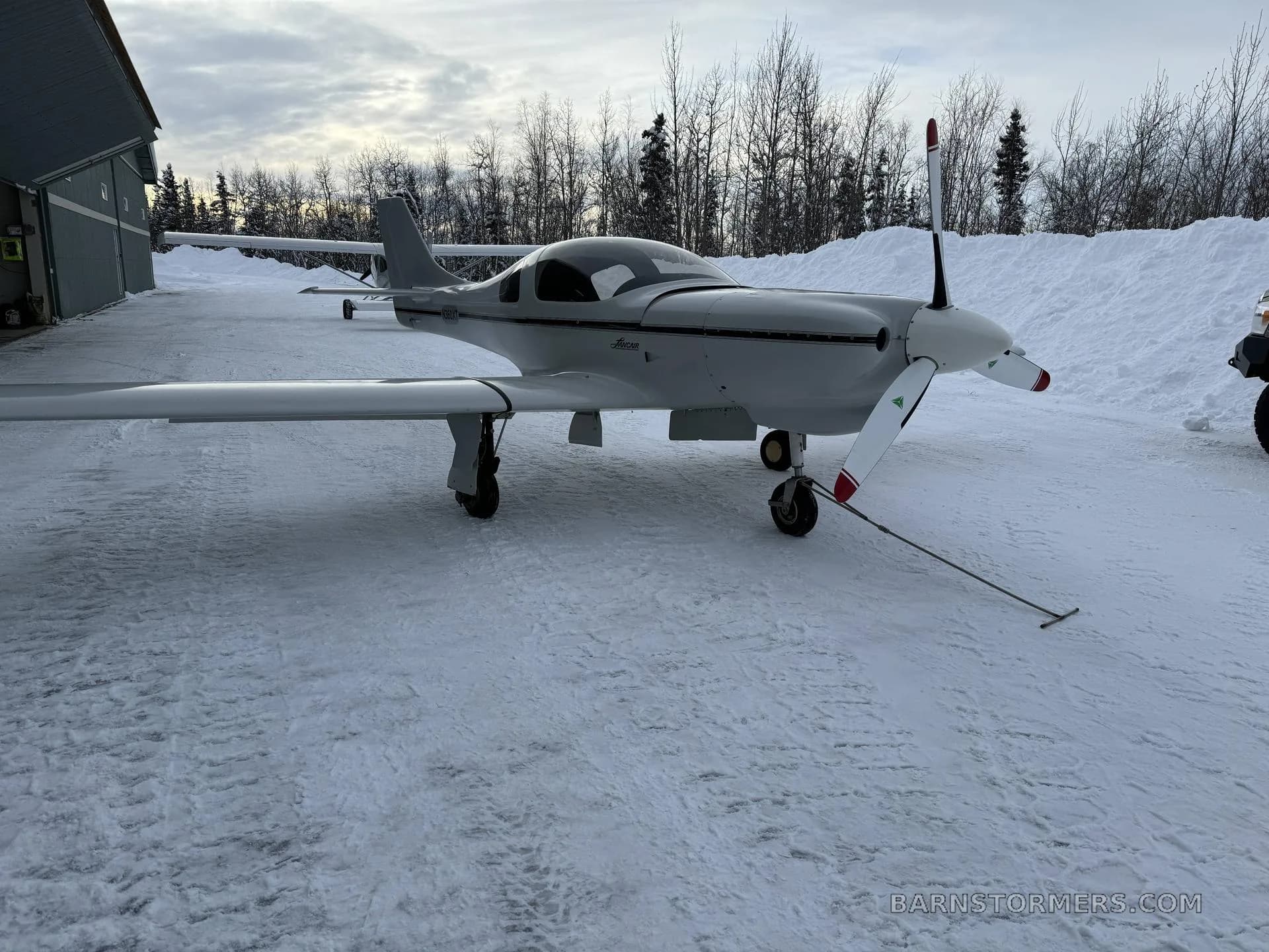 A white Lancair Legacy aircraft with a black stripe is parked on snowy ground next to a hangar, with trees in the background.