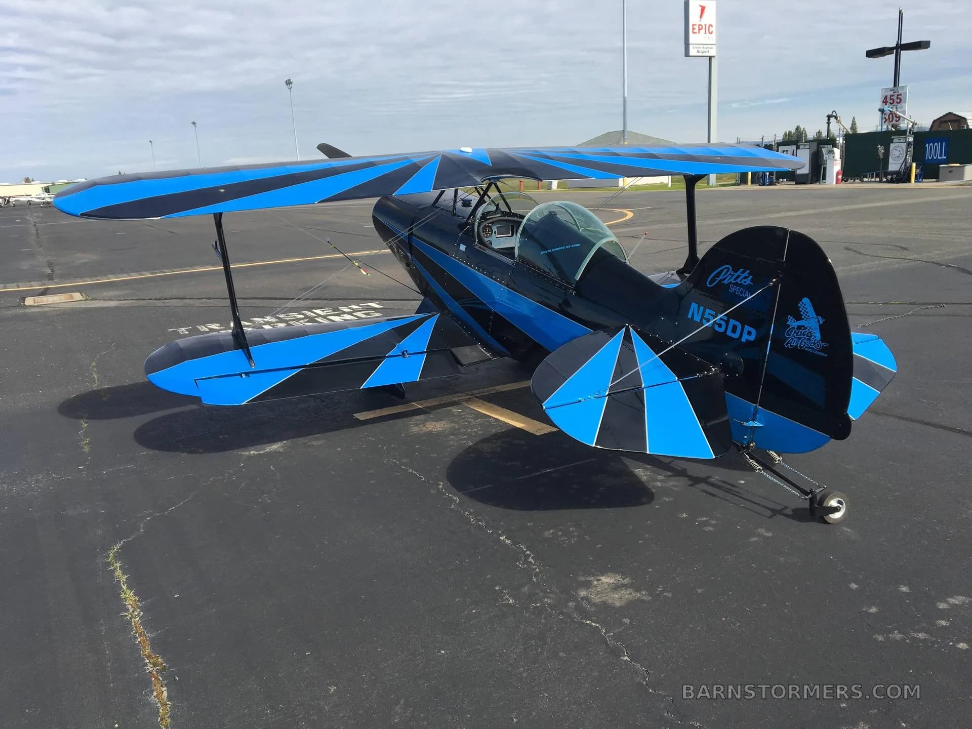 A black and blue Pitts Special biplane, tail number N55DP, is parked on an airfield tarmac with a fuel station in the background.
