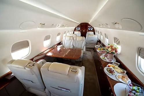 The luxurious cabin interior of a private jet with cream leather seats, a polished wooden table, and catering setup.