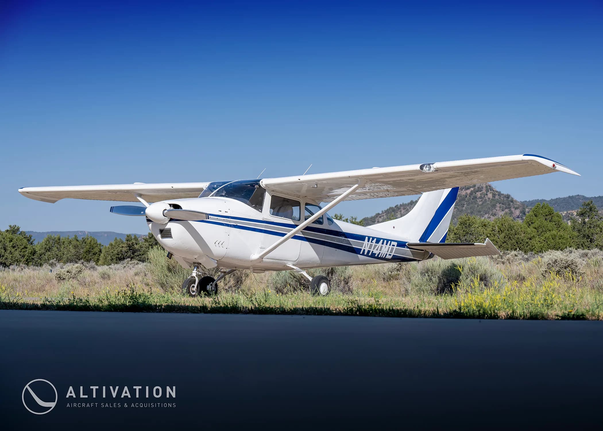 A white Cessna 182 with blue and grey stripes, registration N74MD, viewed from a low angle on a tarmac with mountains in the distance.