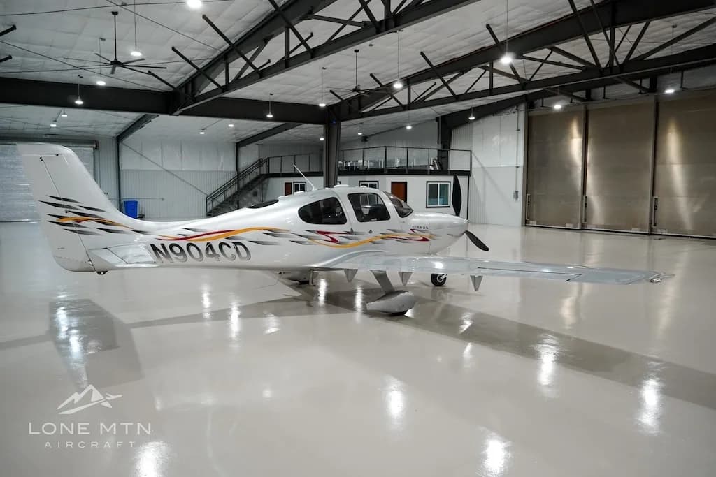 A silver Cirrus aircraft with red and yellow accent stripes, registration N904CD, parked in a hangar.