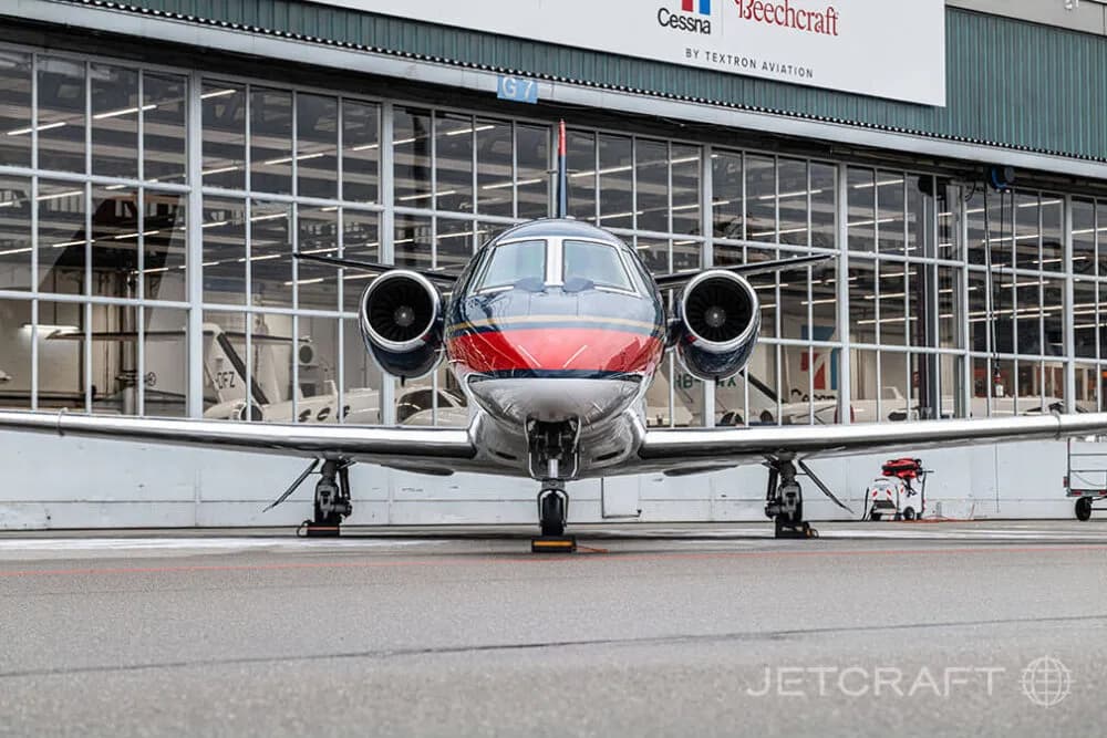 A dark blue, red, and white business jet parked head-on on an asphalt apron in front of a hangar.