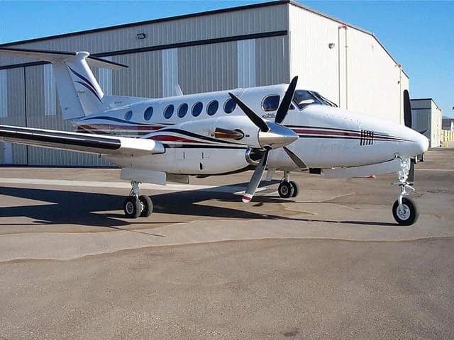 A white twin-engine turboprop aircraft with red and blue stripes parked on an tarmac in front of a hangar.