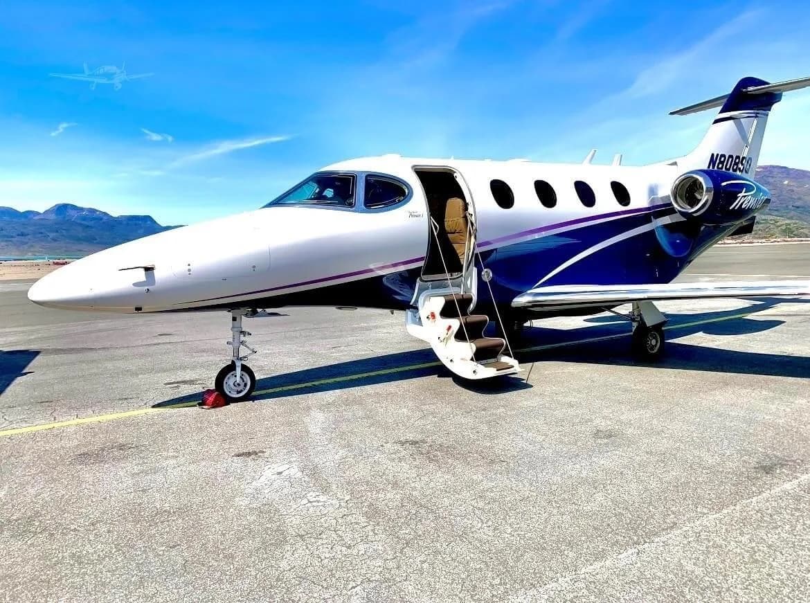 A white business jet with blue and purple stripes, identified as a Premier I, parked on an airfield with its main cabin door open and stairs extended, against a backdrop of mountains and a clear blue sky.
