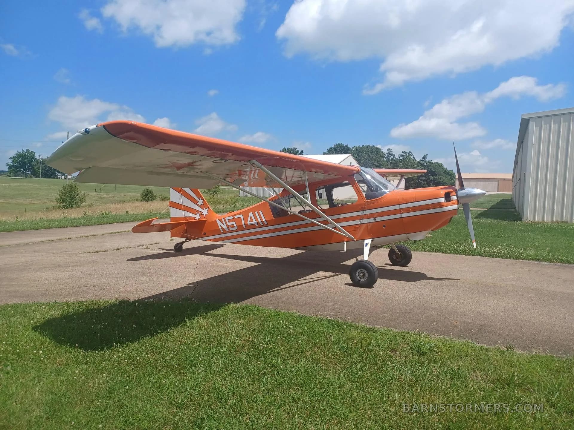 An orange and white high-wing monoplane, registration N57411, parked on a tarmac with a hangar in the background under a blue sky.