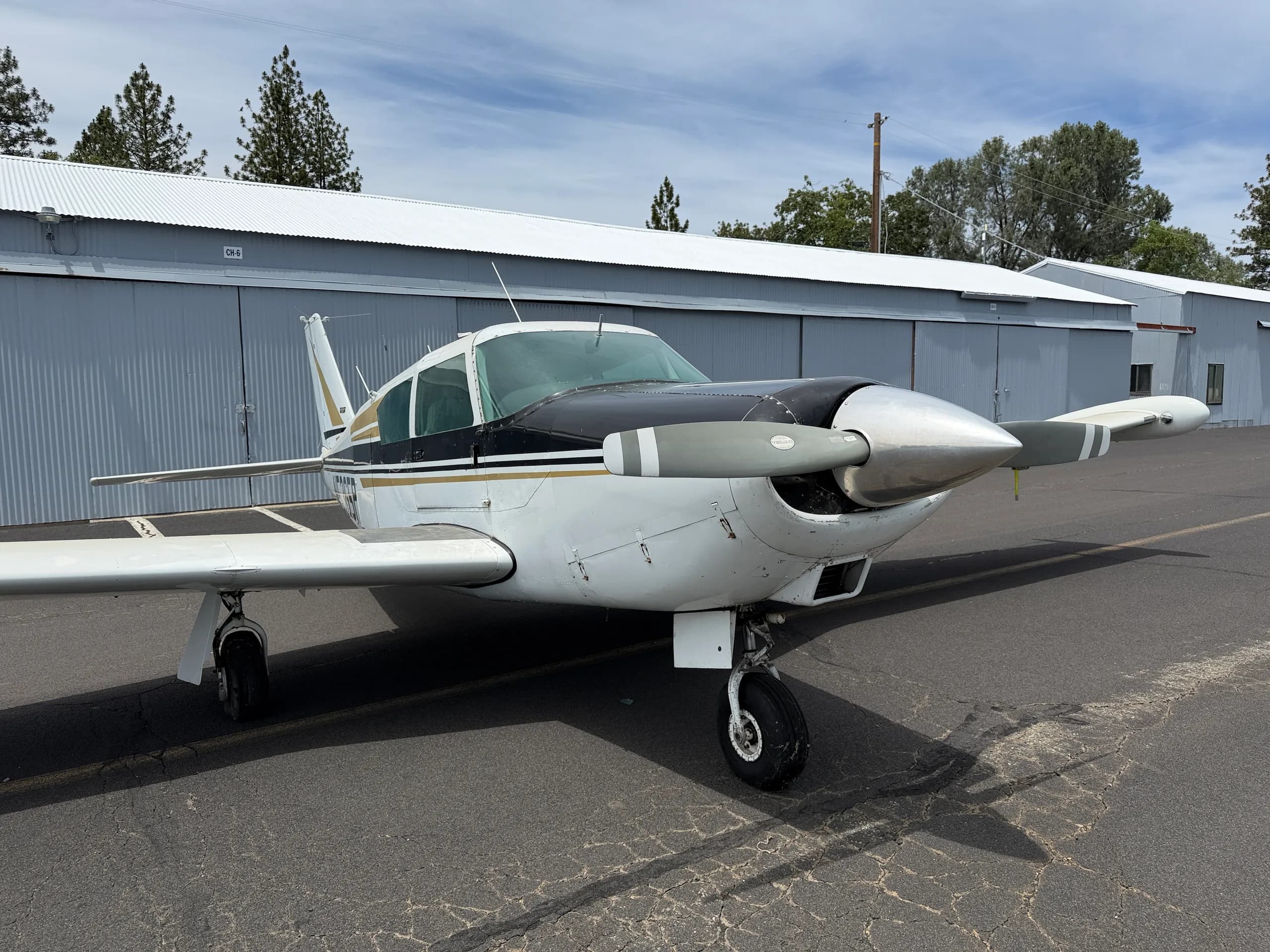 A white single-engine propeller aircraft with black and gold stripes is parked on an asphalt ramp in front of hangars.