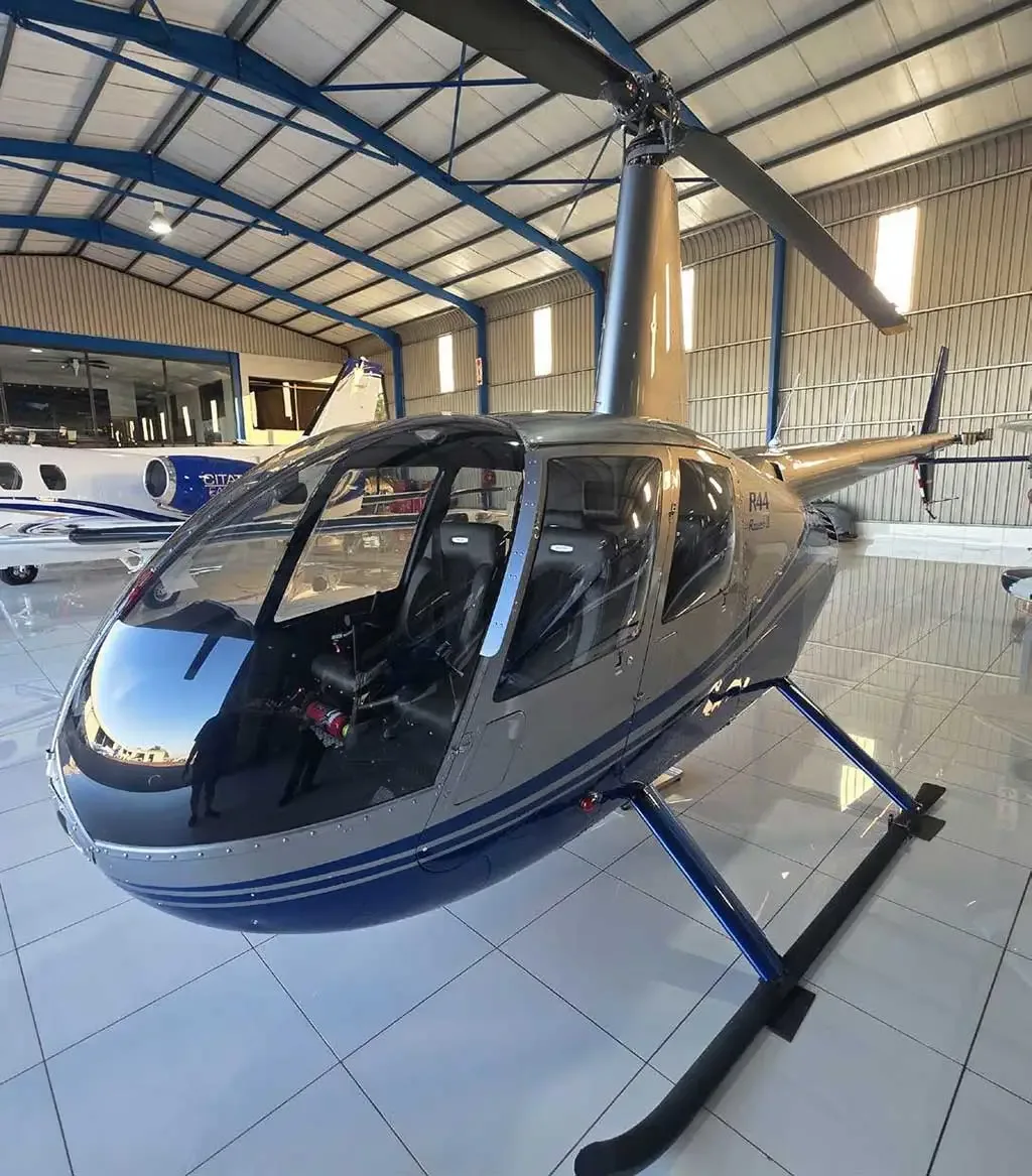 A three-quarter front view of a silver and blue Robinson R44 Raven II helicopter with its doors open in a hangar.