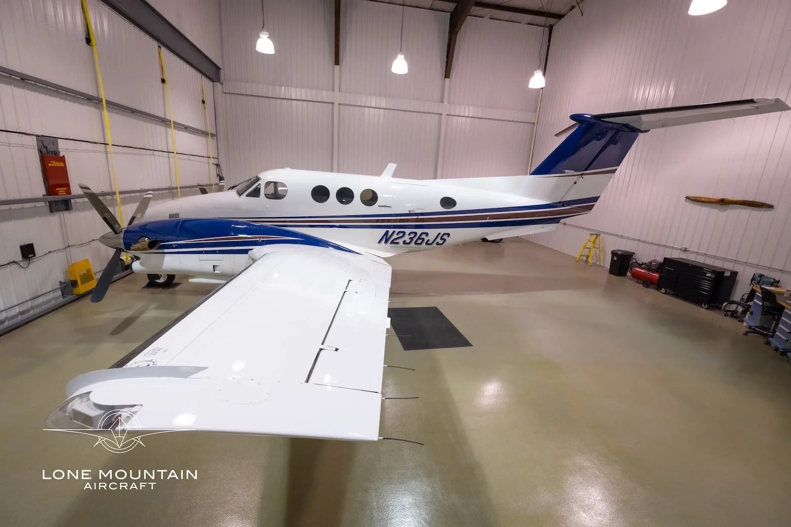 A white and blue twin-engine turboprop aircraft, N236JS, is viewed from the front-right in a hangar.