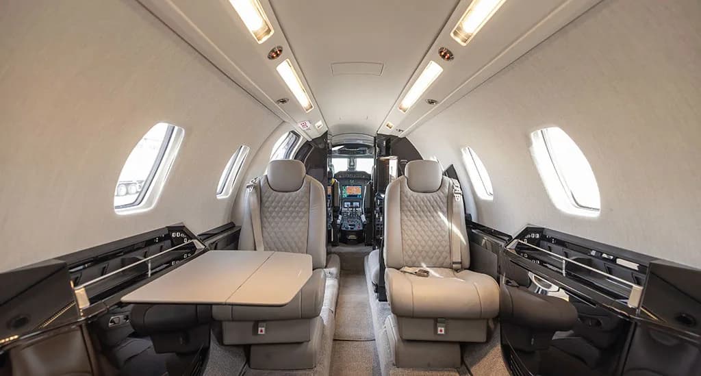 The interior cabin of a private jet featuring light gray quilted leather seats, a deployed table, and dark wood accents.