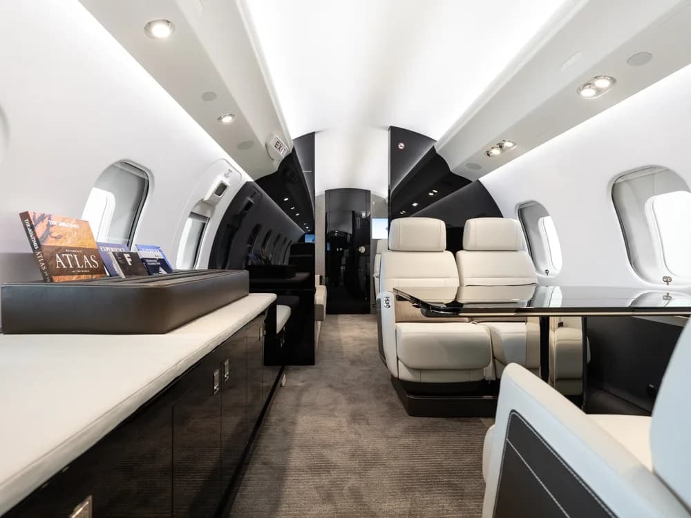 A wide view of a private jet's luxurious cabin interior, featuring light grey seats, a dark console with books, and large windows.