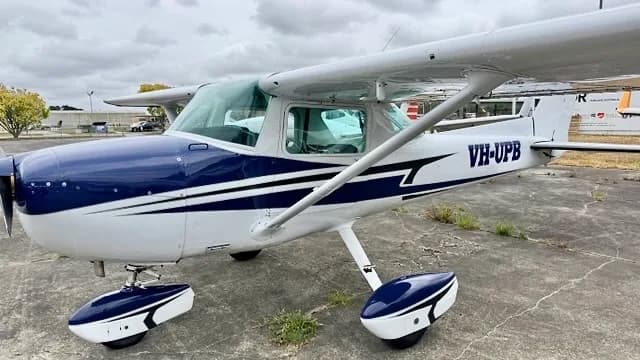 A white and blue single-engine propeller aircraft, registration VH-UPB, parked on an asphalt surface under a cloudy sky.