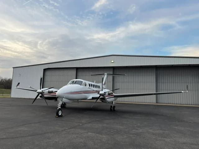 A white Beechcraft King Air with red and black stripes parked on an tarmac in front of a hangar, viewed from the front-left.
