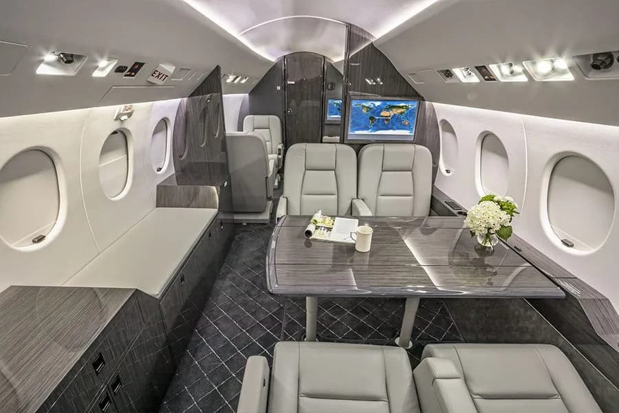 The spacious cabin of a private jet with a large table, grey seats, and a light-colored bench.
