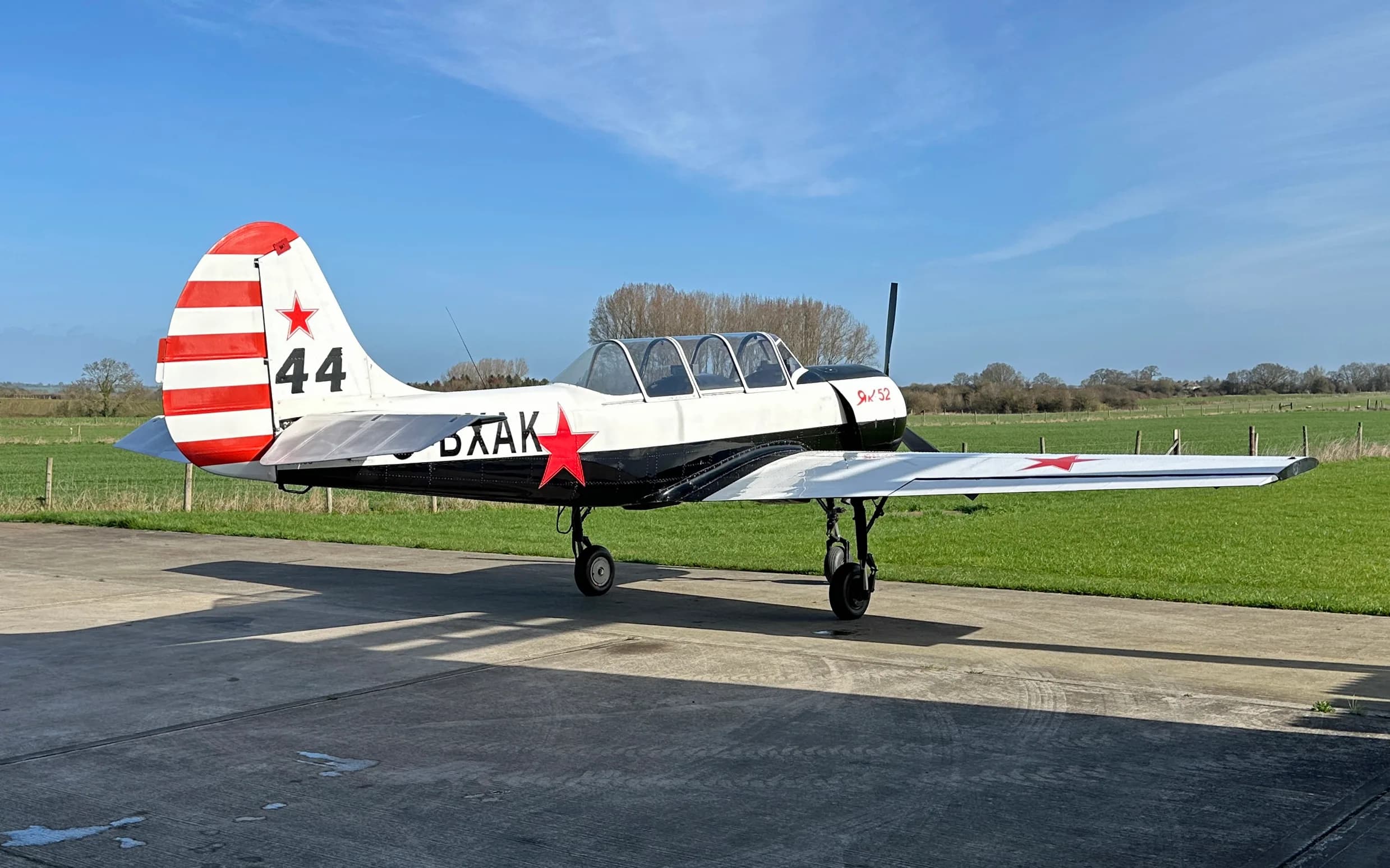 A white and black Yak-52 aircraft with red stars and tail stripes, parked on a concrete pad next to a grassy field.