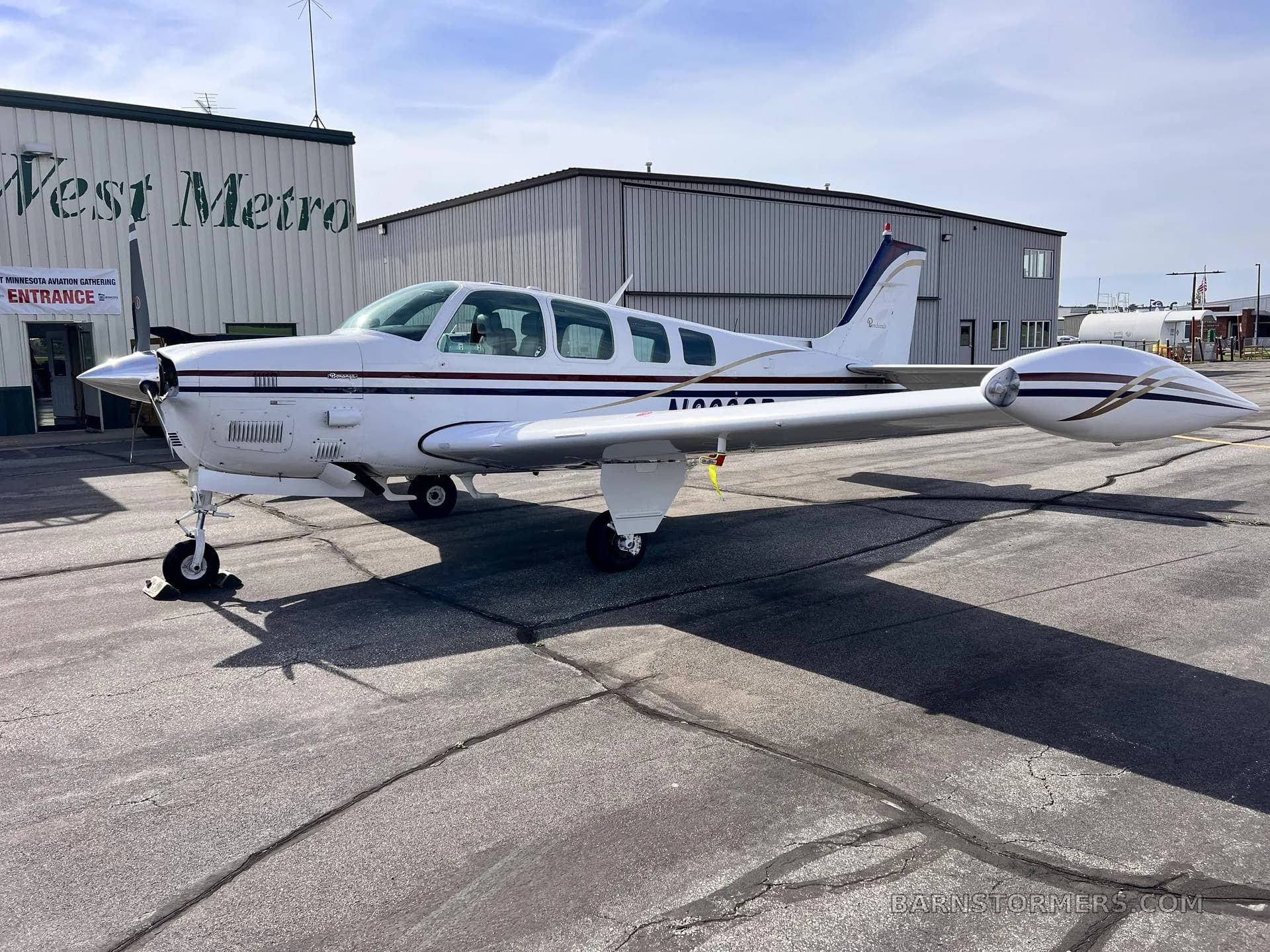 A white Beechcraft Bonanza with dark blue and gold stripes is parked on a tarmac next to a hangar.