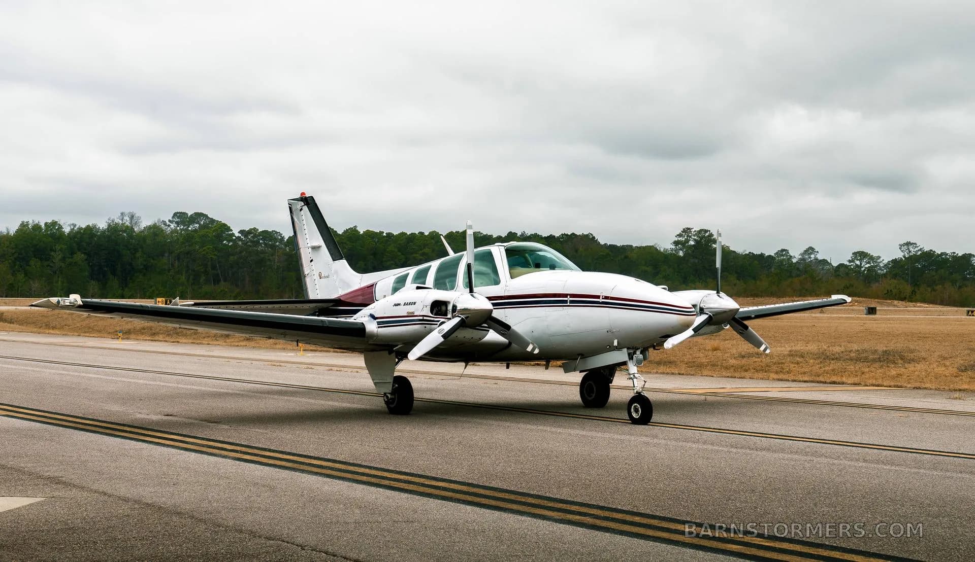 A white twin-engine Beechcraft Baron N158TB with maroon and blue stripes is seen from the front-right on a taxiway under a cloudy sky.